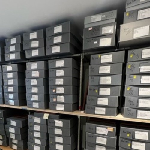 Large grey acid free boxes stacked on shelves with white labels specifying the artifact contents inside.