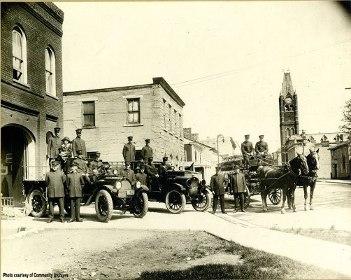 Firefighters with horses and new pumper trucks in front of lower fire hall with City Hall in the background, circa 1922