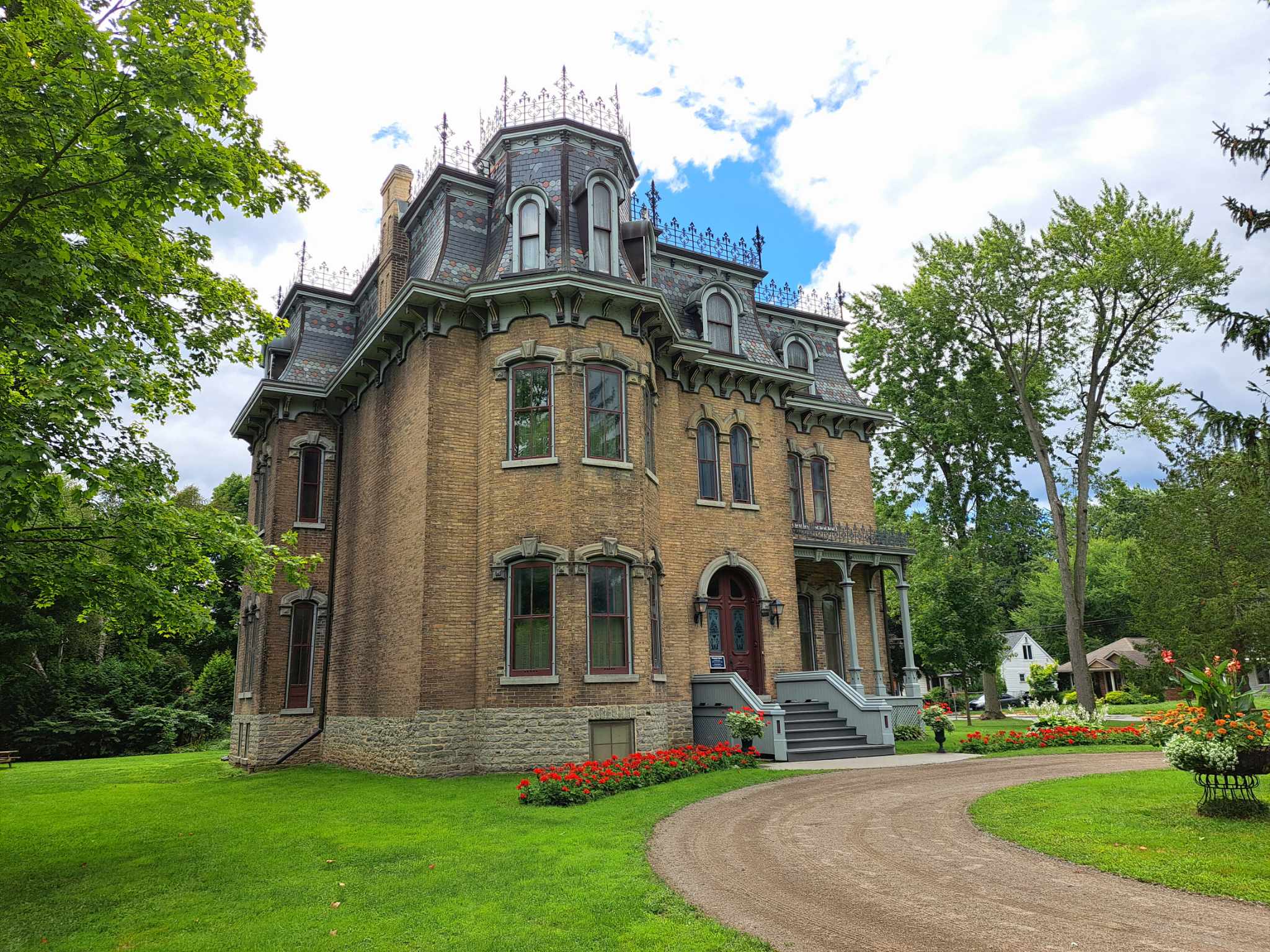 The facade of a large brick home with a mansard roofline. The flower beds in front of the house feature red flowers and part of the circular gravel driveway is visible. 