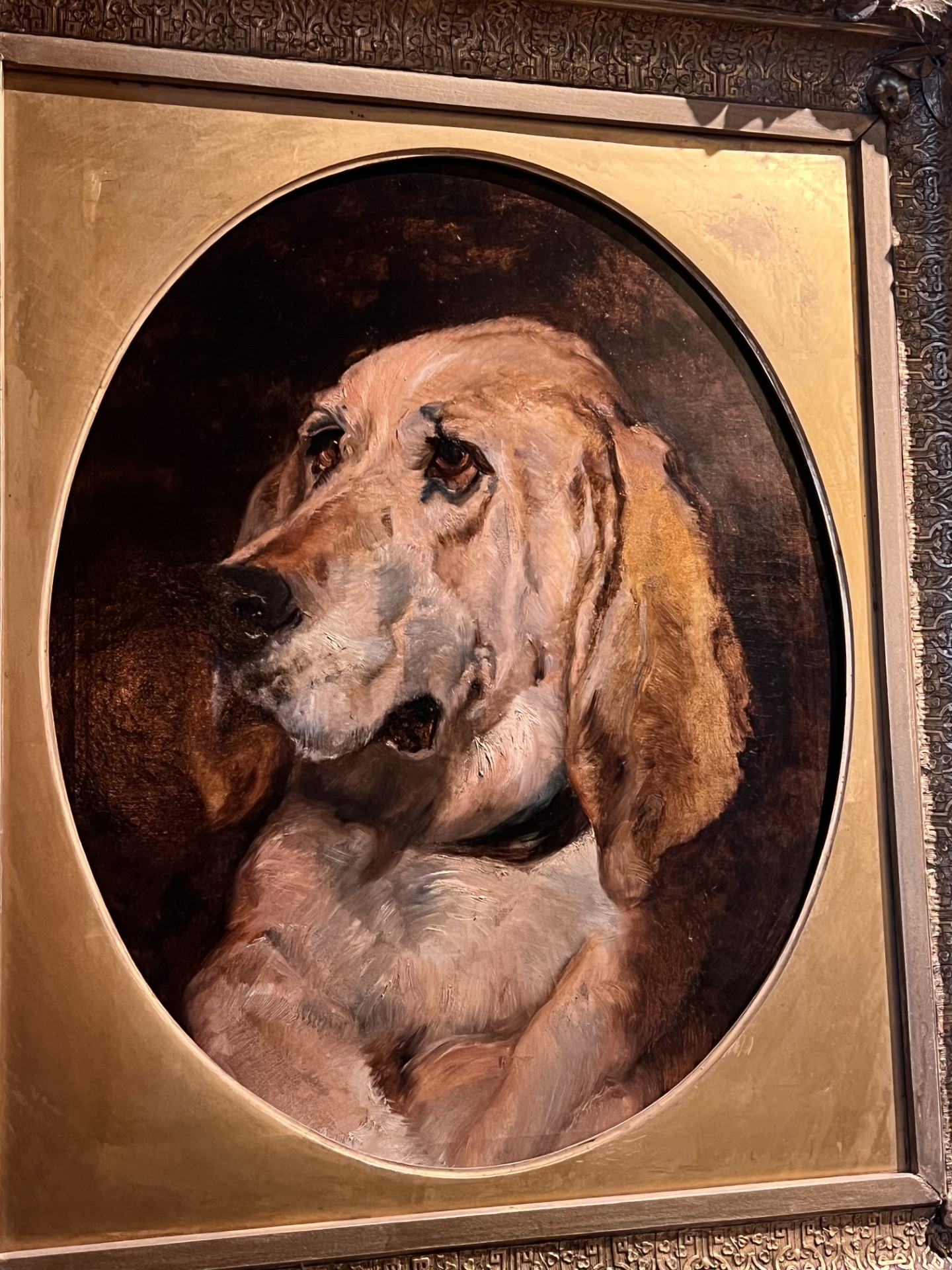 An oil painting study of a blood hound.  It has an oval mat and an elaborate gold frame. This copy is attributed to Bertram Couldery. It is a study of an original painting by Sir Edwin Landseer called Dignity and Impudence. 
