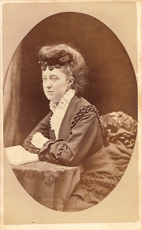 A woman seated for a portrait wearing a dark dress with a white ruffle along the neckline. She has an elaborate updo hairstyle. 