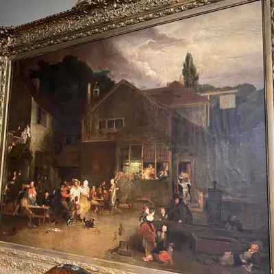 An oil painting in an elaborate gold frame. The scene depicts a group of people gathered outside of a tavern.  Attributed to Bertram Couldery this is a copy of The Village Holiday by Sir David Wilkie