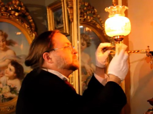 A man wearing a black coat and white gloves adjusts a wall light.