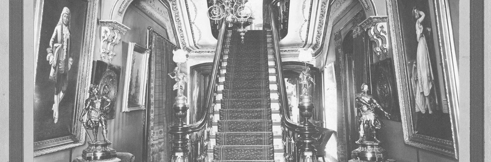 An elaborate central hall of a Victorian home featuring a suspended column staircase which is flanked by statues and large figural paintings.