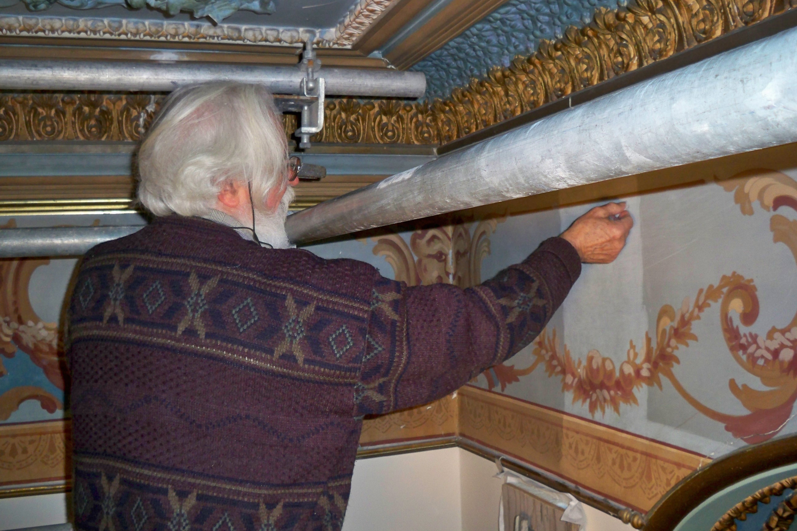 Conservation in action at Glanmore. A man with white hair and a beard uses a moldable eraser to clean the delicate hand-painted finishes on the wall of Glanmore's drawing room. 