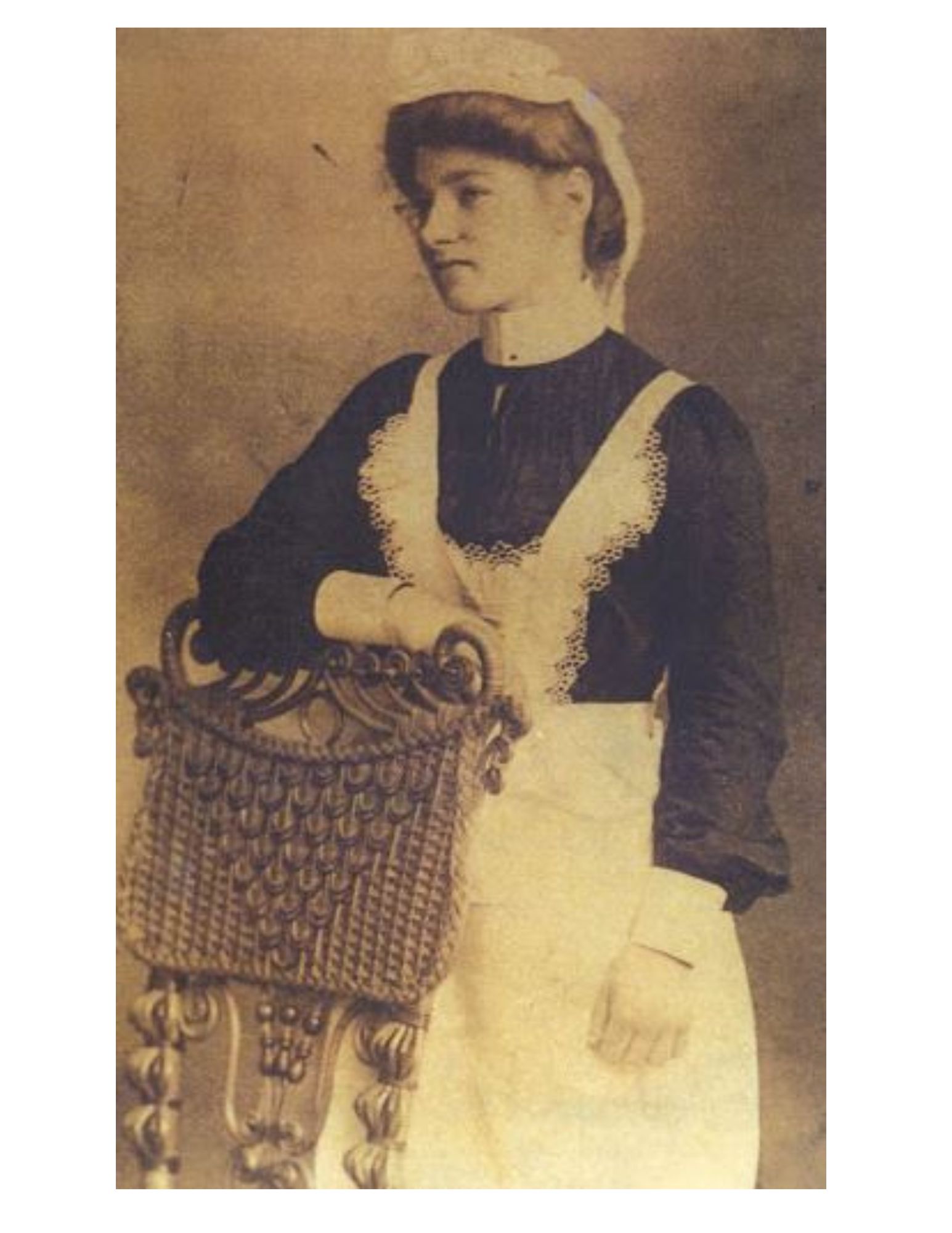 A sepia toned portrait of a young woman, circa 1901, standing in front of a wicker chair. She is wearing a black dress, white apron and white lace bonnet. 
